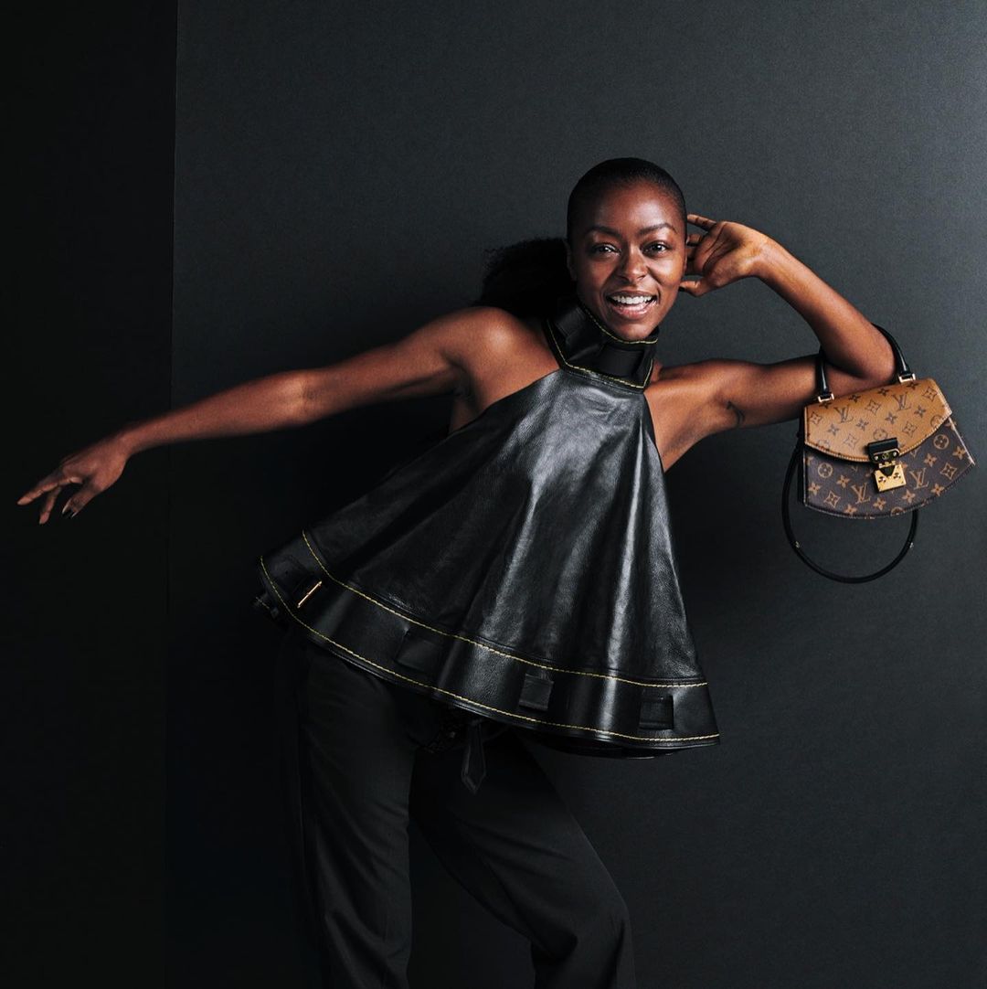 A smiling African-American woman poses in a black, halter-style top with a brown Louis Vuitton handbag against a dark background.