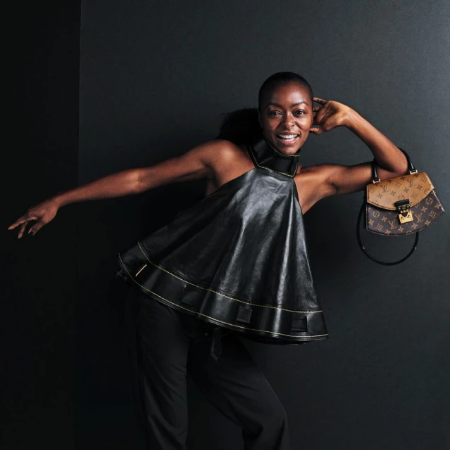 A smiling African-American woman poses in a black, halter-style top with a brown Louis Vuitton handbag against a dark background.