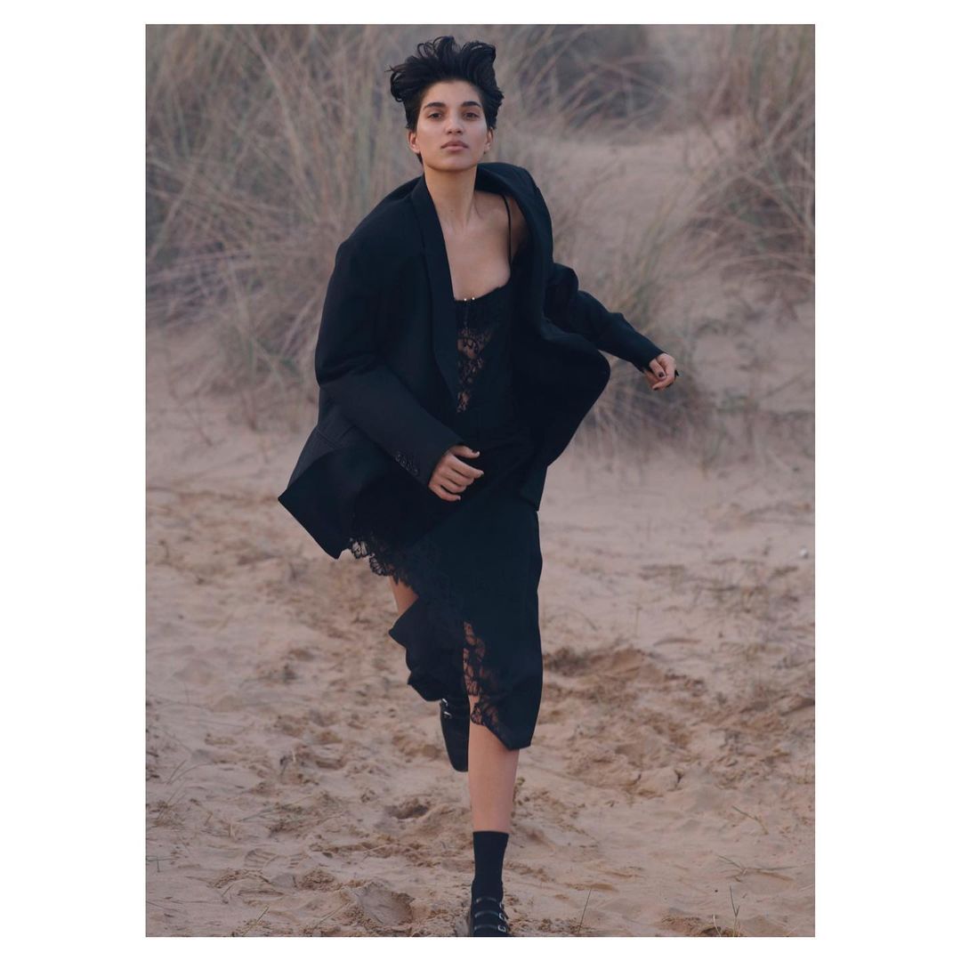 Woman in dark blazer and black lace dress running across a sandy coastal landscape with tall dried grass.