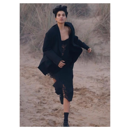Woman in dark blazer and black lace dress running across a sandy coastal landscape with tall dried grass.