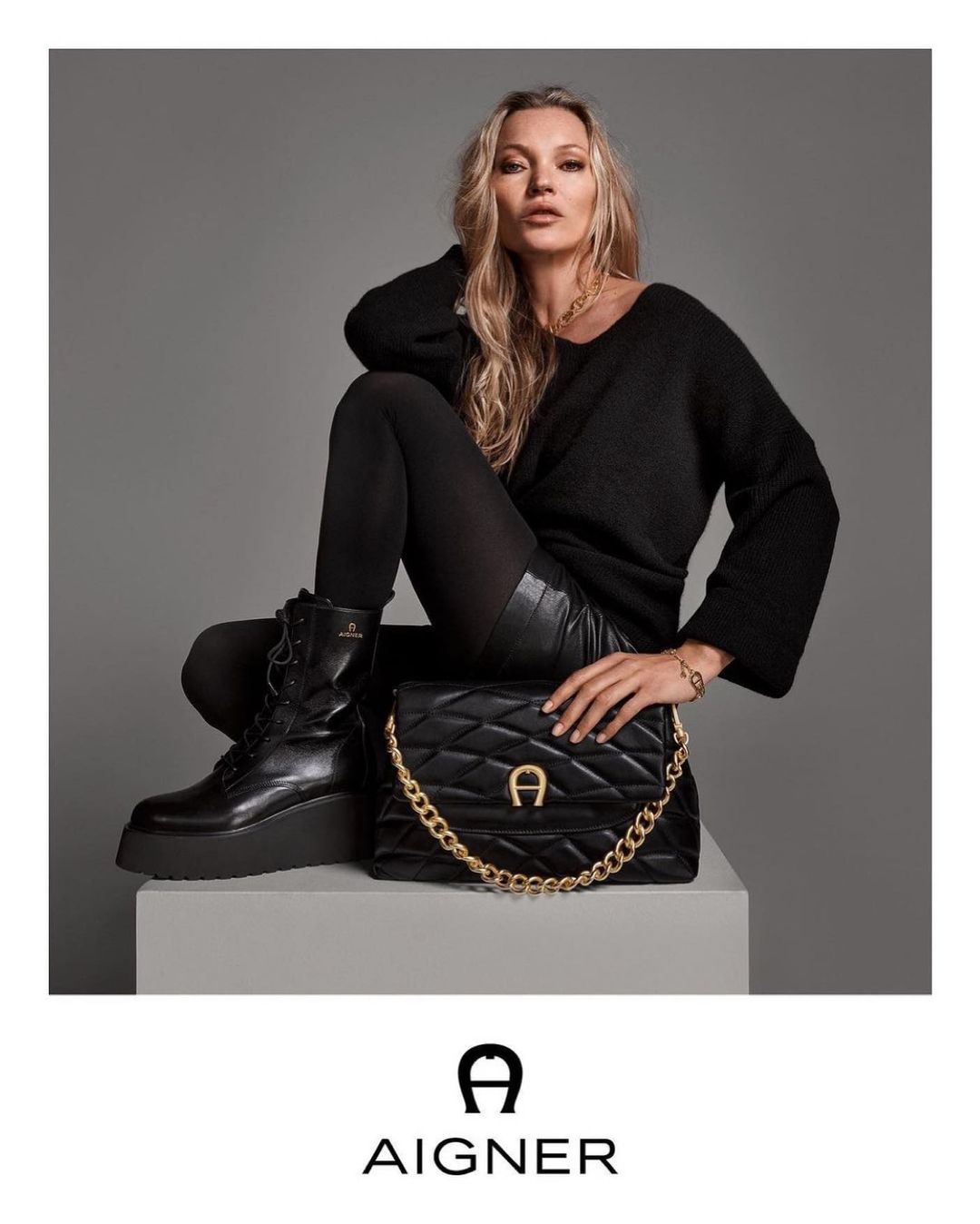 Kate Moss sits in a fashion shot for Aigner, wearing a black sweater, leather skirt, tights, and boots, holding a black quilted purse.