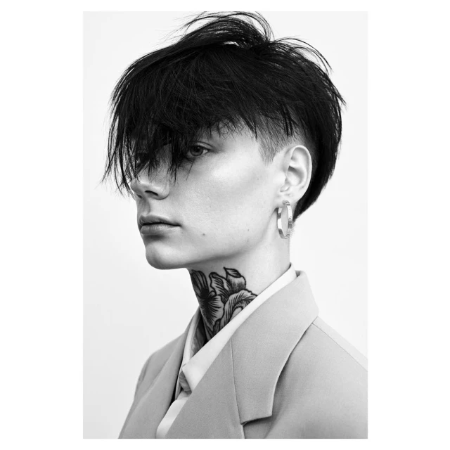 Black-and-white portrait of a model wearing a blazer, featuring a neck tattoo and hoop earrings, captured in a studio setting.