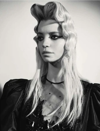 Portrait of a woman with dramatic hairstyle and dark eye makeup wearing a black dress against a plain background