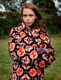 Portrait of a young woman with long hair wearing a patterned fuzzy coat outdoors in a grassy field with trees.