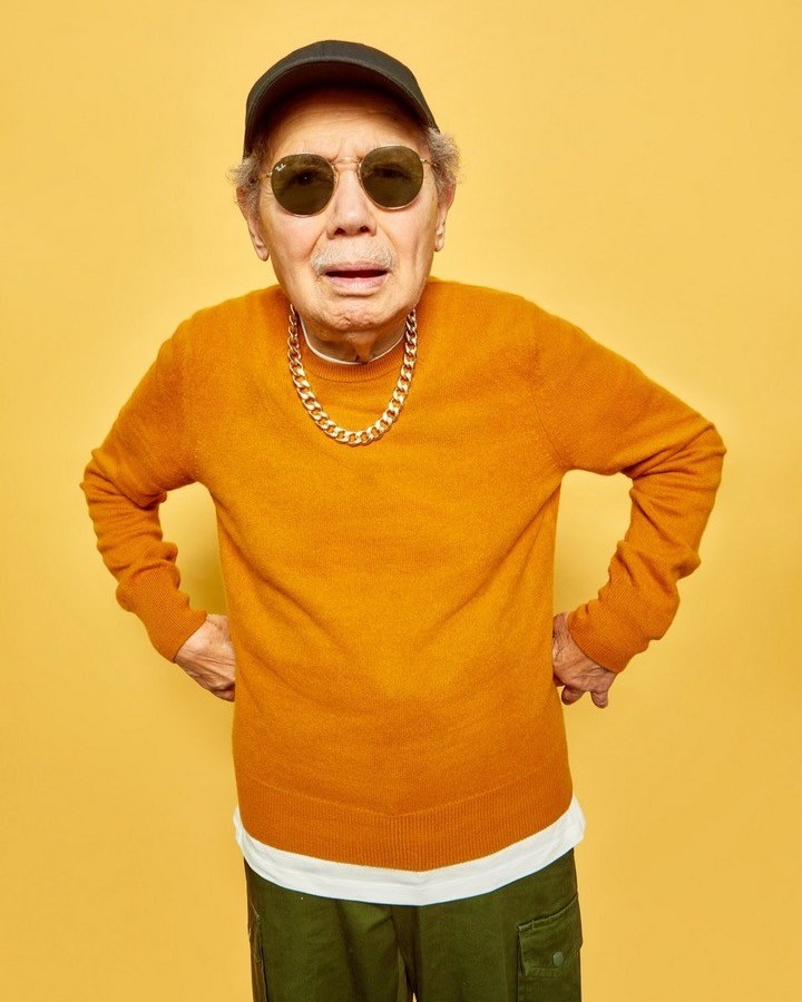 Elderly man in orange sweater, black baseball cap, and sunglasses posing against a yellow background with hands on hips.