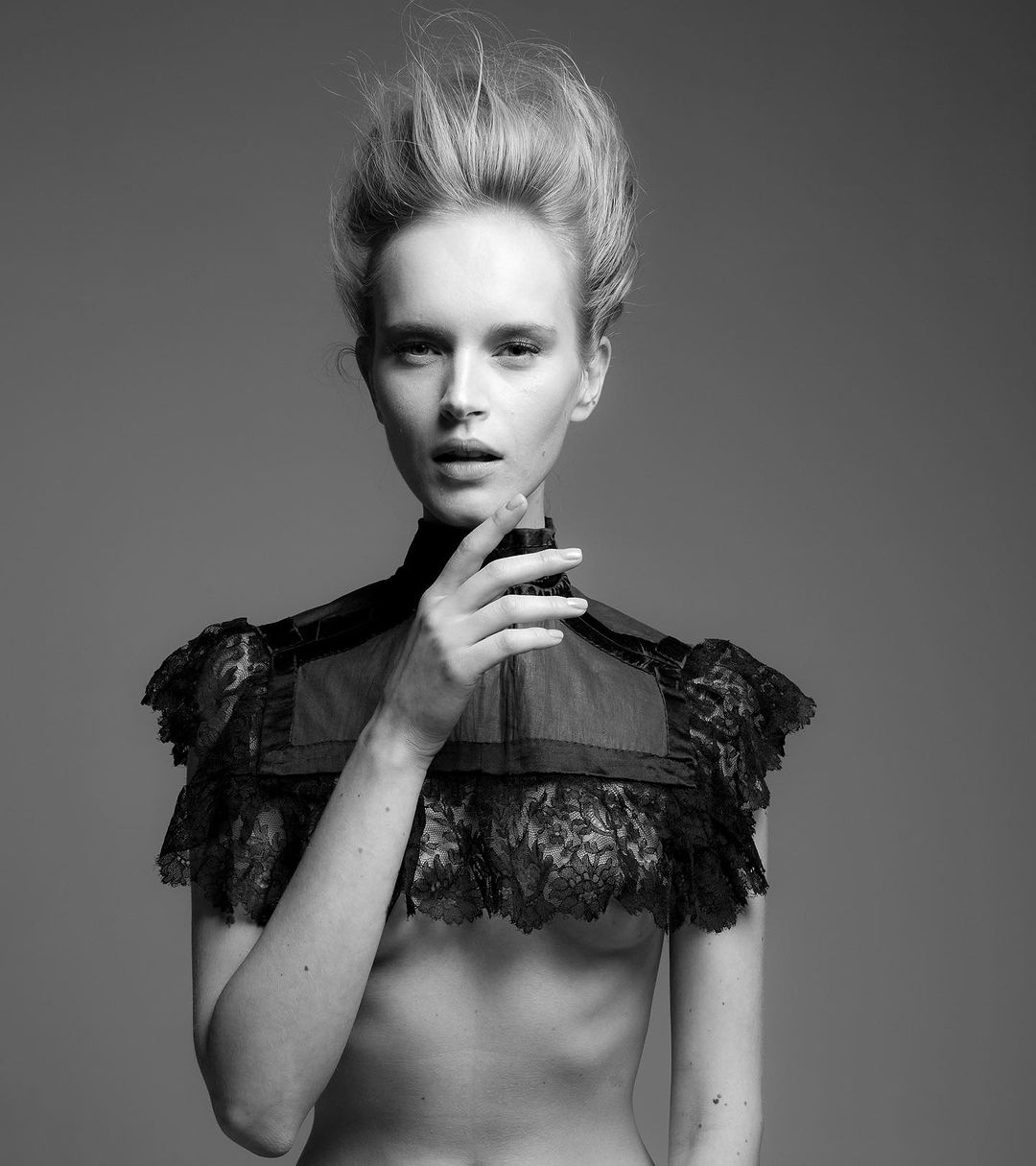 Black and white portrait of a woman wearing a lace collar with her hand near her face, against a neutral gray backdrop in a studio.