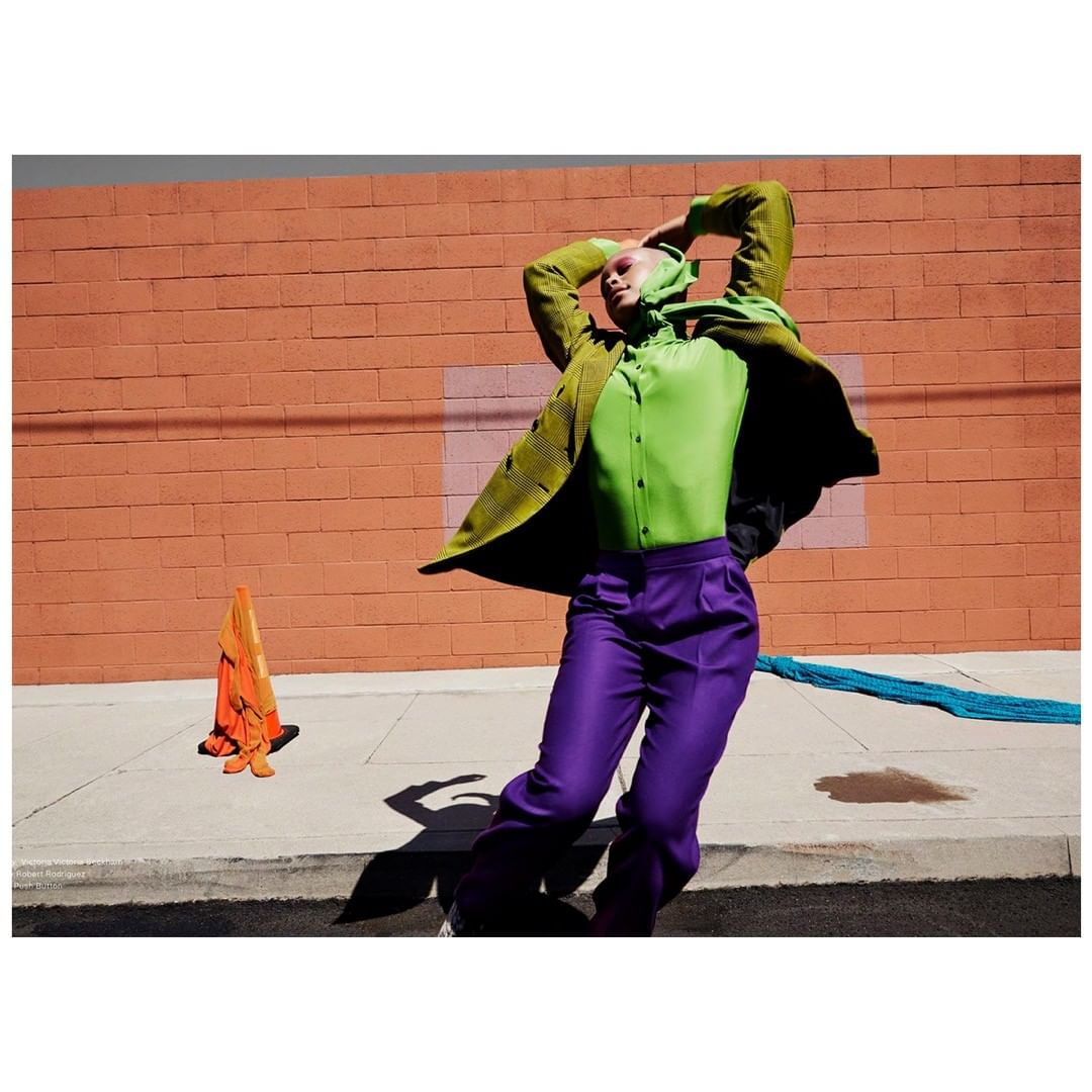 A stylish person with green top and purple pants poses outdoors in front of an orange brick wall, with a traffic cone nearby.