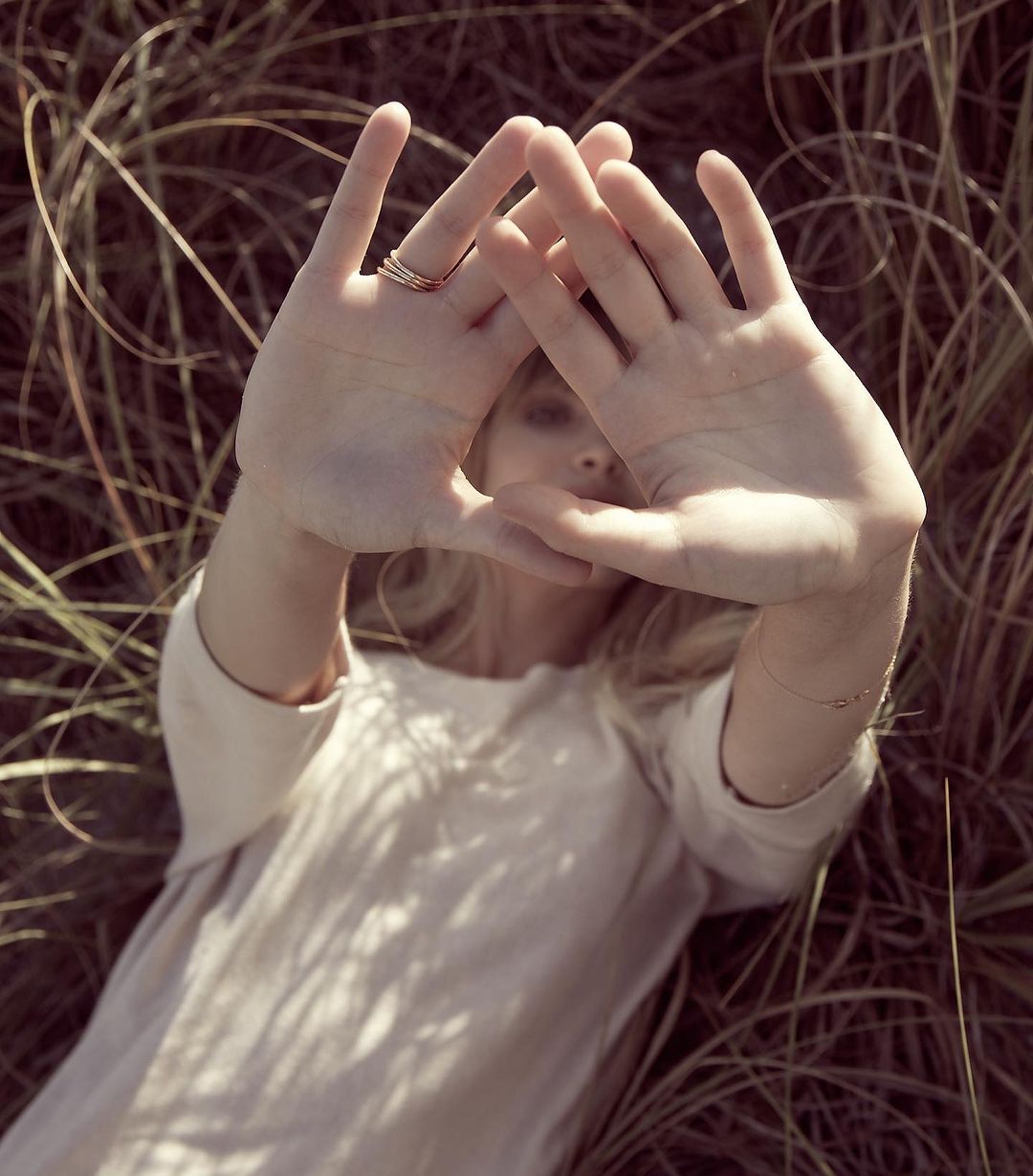 Blonde woman lies in a field of tall grass with hands raised in a triangle shape, partially obscuring her face, in soft focus and light.