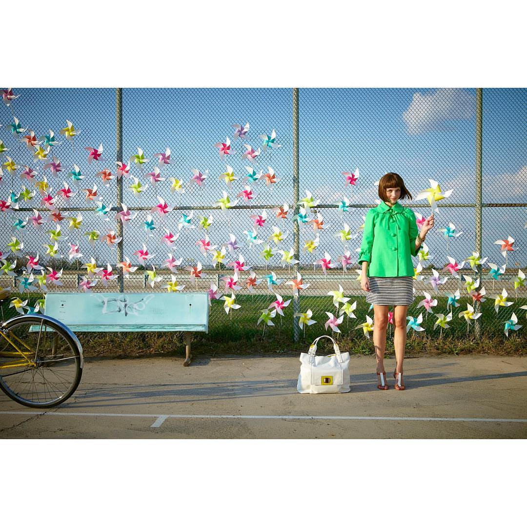Woman in green jacket stands with colorful pinwheels attached to a fence, next to a bench, a bicycle, and a tote bag outdoors.