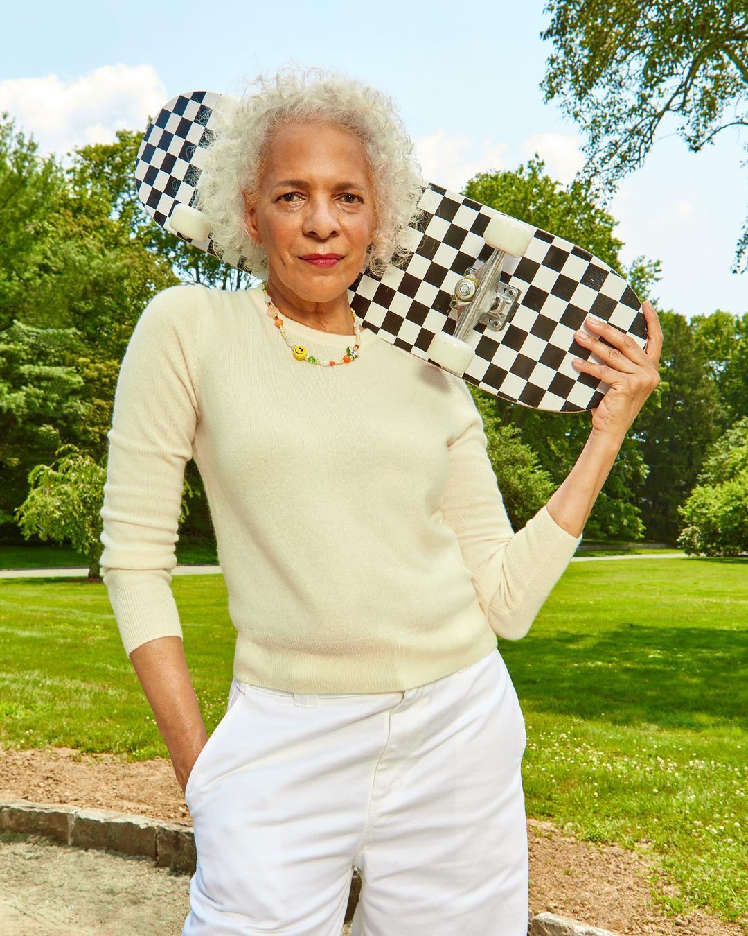 Stylish mature woman with curly white hair posing outdoors with a black and white checkered skateboard in a sunny park setting.