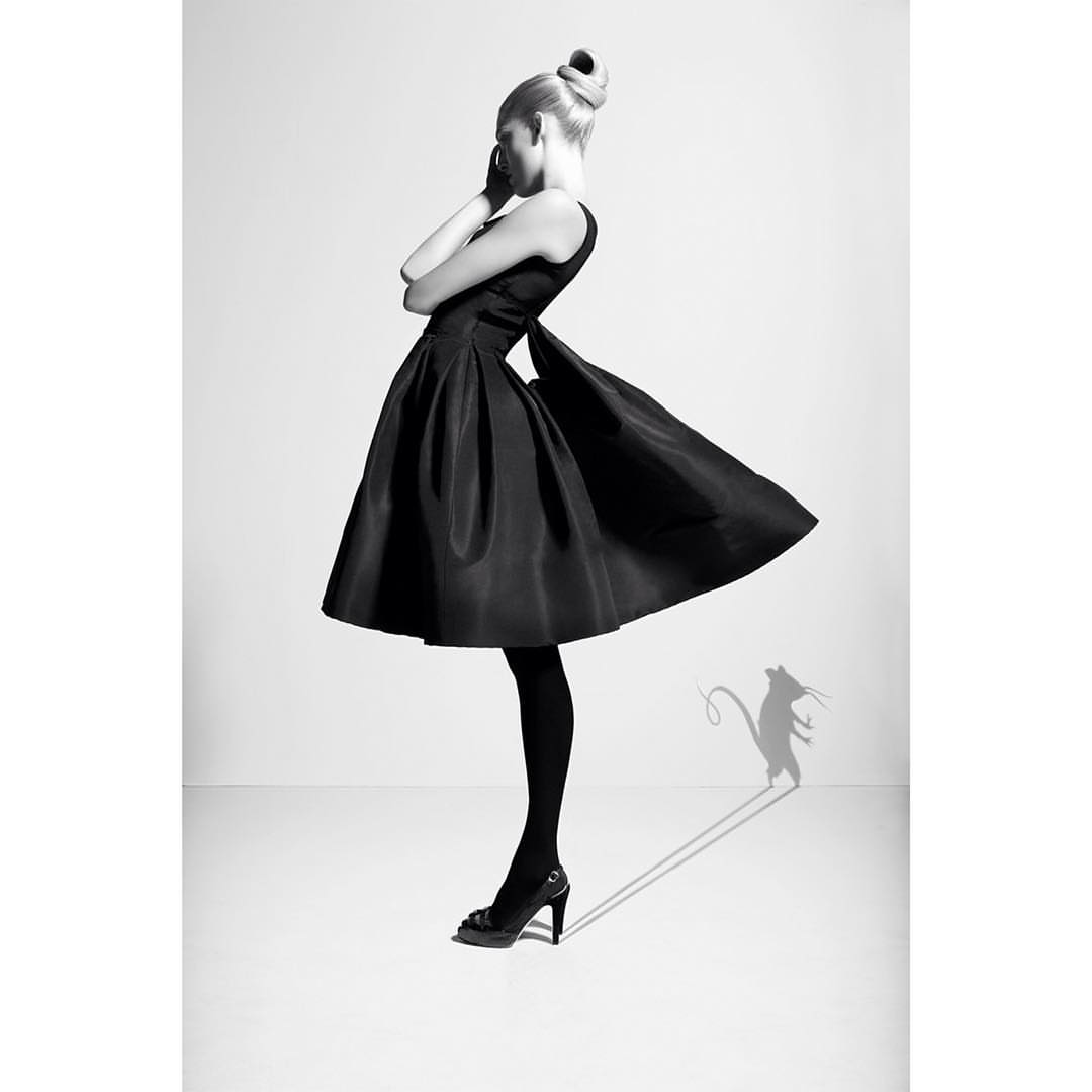 Elegant woman in black dress and high heels poses in a photo studio with a rat shadow in a monochrome fashion shot.