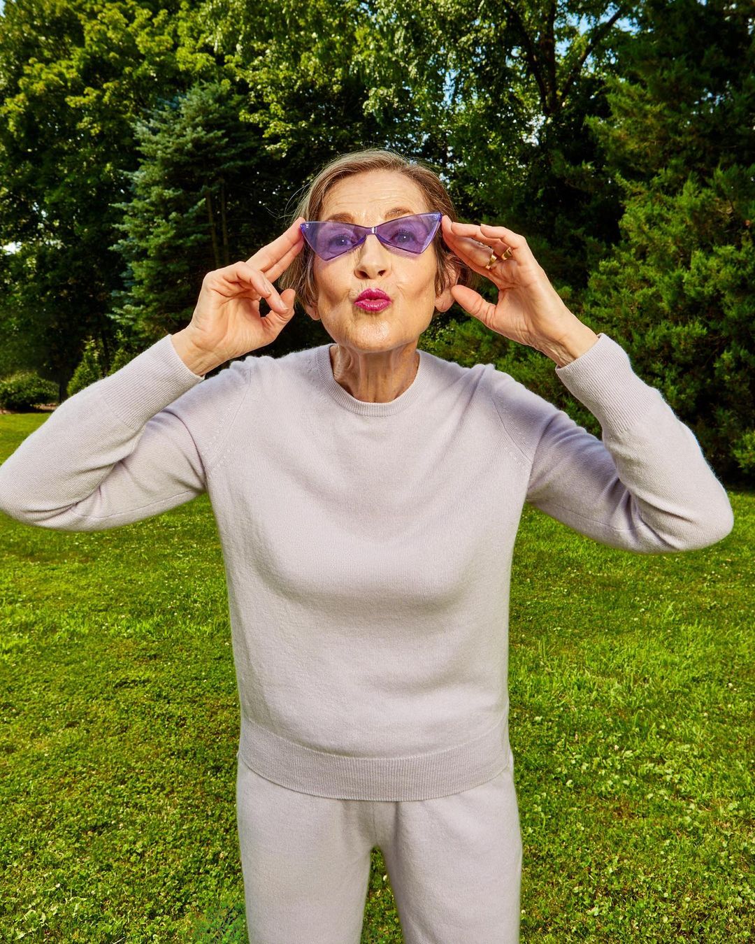 Elderly woman outdoors in lavender outfit and sunglasses, playfully posing in front of a green lawn and trees on a sunny day.
