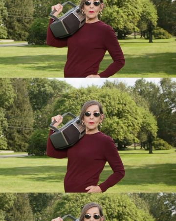 Mature woman in sunglasses carries a vintage boombox over her shoulder, standing on a grassy lawn with trees in the background.