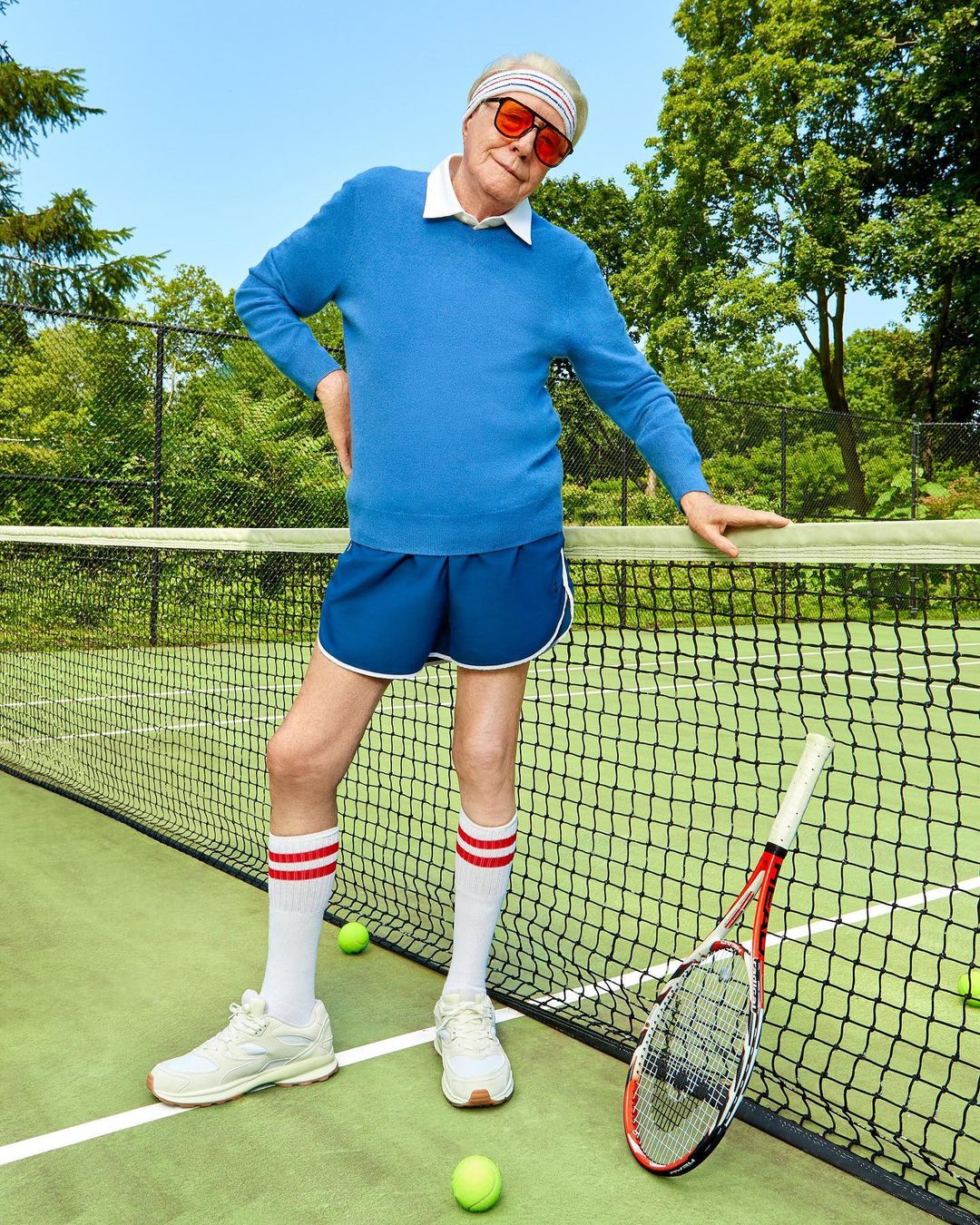 Older adult poses with tennis racket and balls on a green court, wearing a blue sweater, shorts, headband, socks, and tennis shoes.