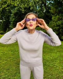 Elderly woman wearing purple sunglasses and a light purple sweater outdoors in a grassy park setting