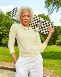Elderly woman with curly white hair wearing an off-white sweater holding a black and white checkerboard skateboard outdoors in a park.