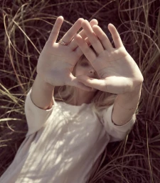 Woman lying amidst tall dry grass holding her hands up to her face forming a triangular frame