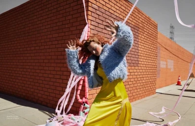 Red-haired model in fuzzy blue jacket and yellow dress posing in front of a red brick building with pink ribbons.
