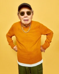 Elderly man wearing amber sunglasses, a gold chain, mustard sweater, and black cap against a yellow background.