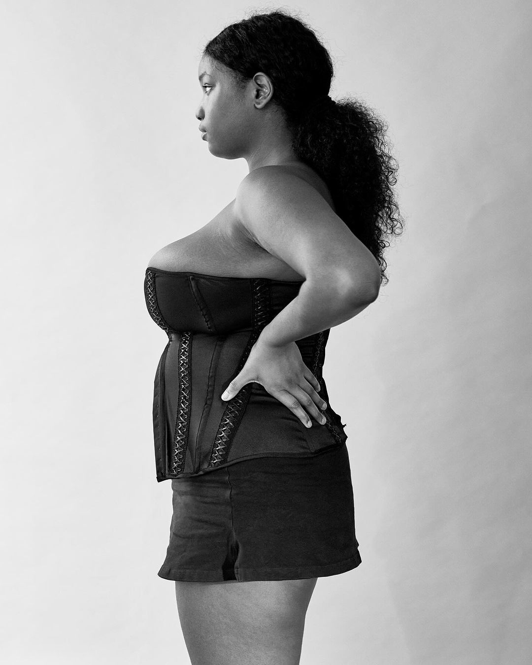 Black and white portrait of an adult woman wearing a corset and shorts, posing indoors with a neutral expression, side view.