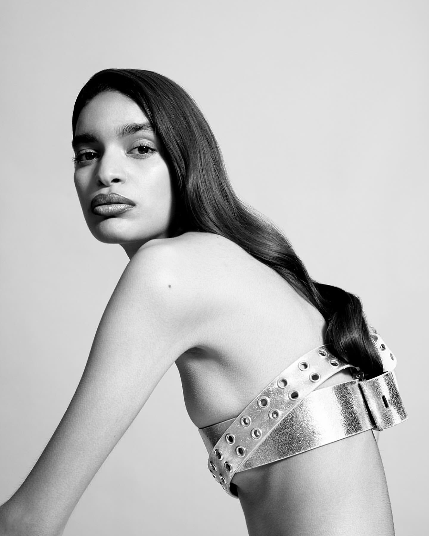 Monochrome portrait of a woman with long dark hair wearing a wide silver belt-like top with grommets against a white background.