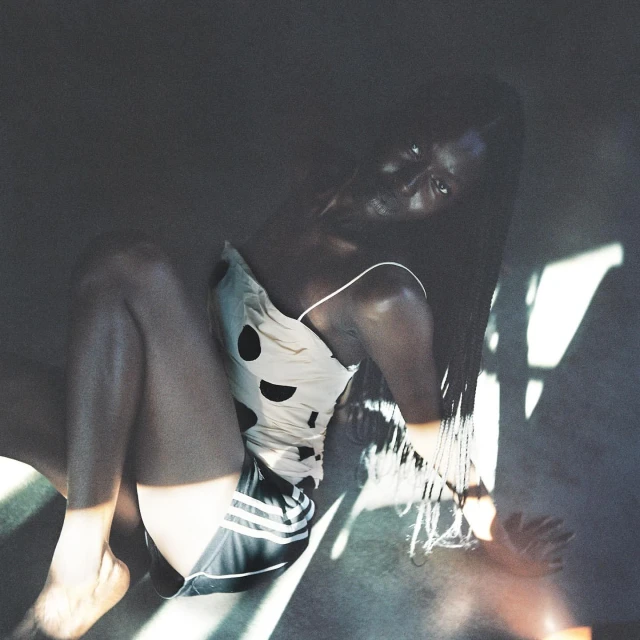 Portrait of a Black woman in a polka-dot top and Adidas shorts sitting on the floor in soft, filtered light with dark tones.