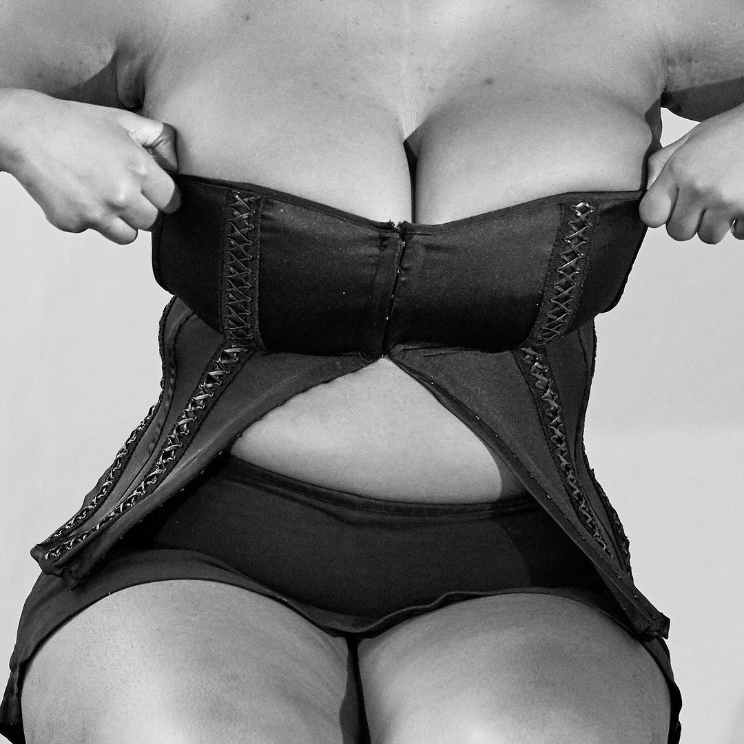 Close-up studio shot in black and white of a curvy woman wearing a corset-style top. Hands are at the upper portion of the garment.