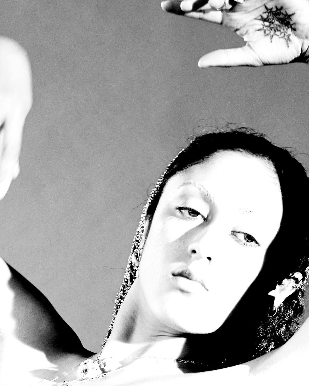 Monochromatic portrait of a woman with decorative hairpiece, arm raised, and a tattooed hand in the background against a grey backdrop.