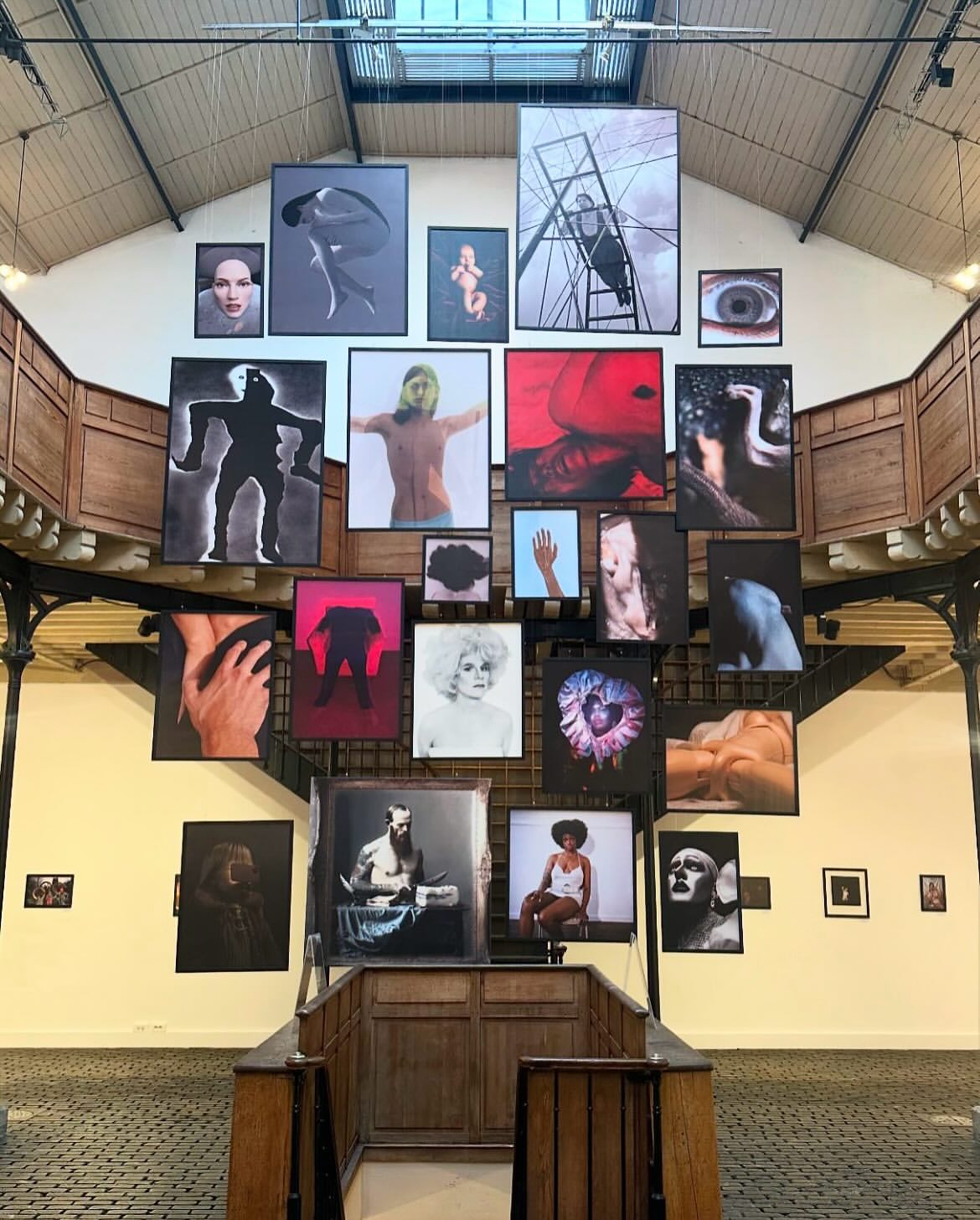 Art exhibition with hanging photographic portraits and figure studies displayed in an indoor gallery space featuring wood and metal architecture.