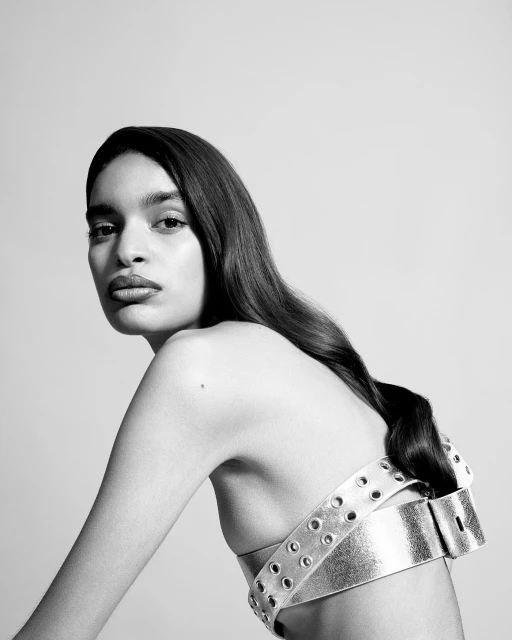 Striking black and white portrait of an adult woman with long dark hair wearing a stylish silver belt as a top, set against a plain background.