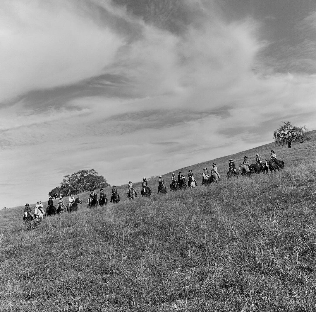 Black and white shot of a group of people on horseback in an open grassy field with scattered trees under a cloudy sky.