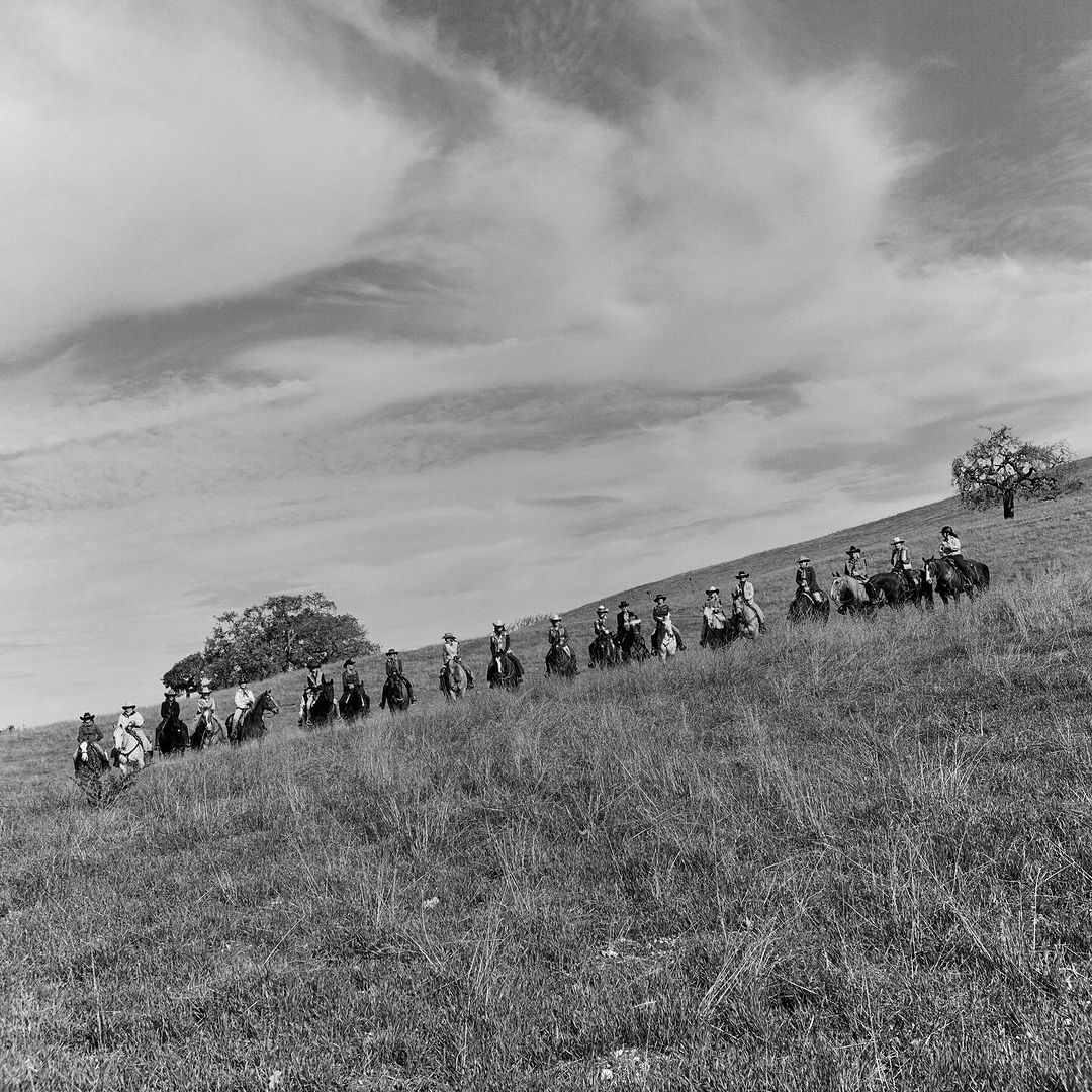 Black and white shot of a group of people riding horses in a line formation across a grassy, sloping hill beneath a cloudy sky.