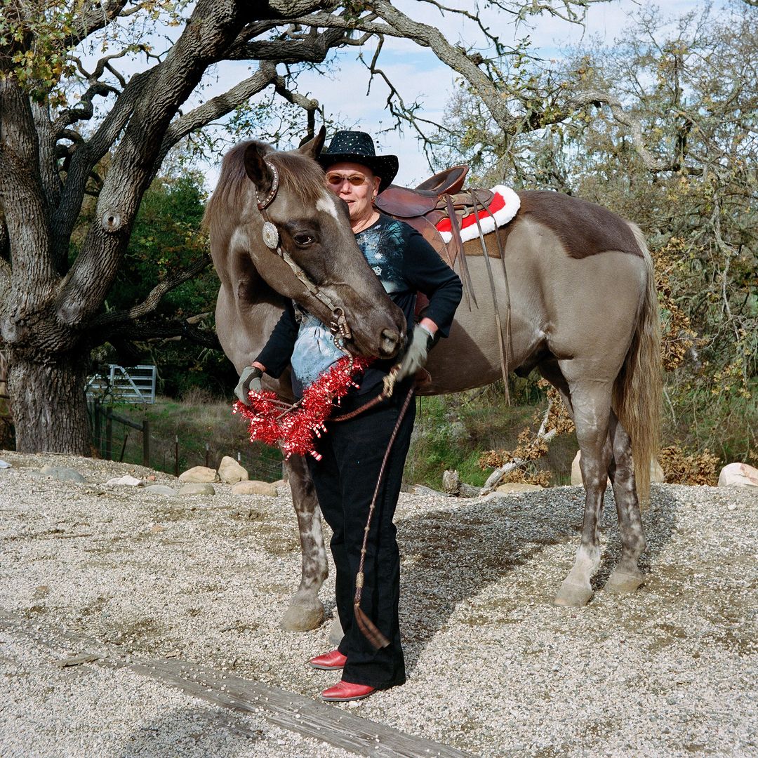 Woman in a cowboy hat poses with a decorated horse outdoors on a gravel surface near trees under a cloudy sky in natural light.