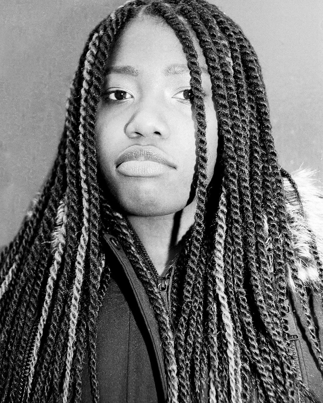 Monochrome portrait of a young woman with long twist braids, fair skin, and a neutral expression against a plain gray backdrop.