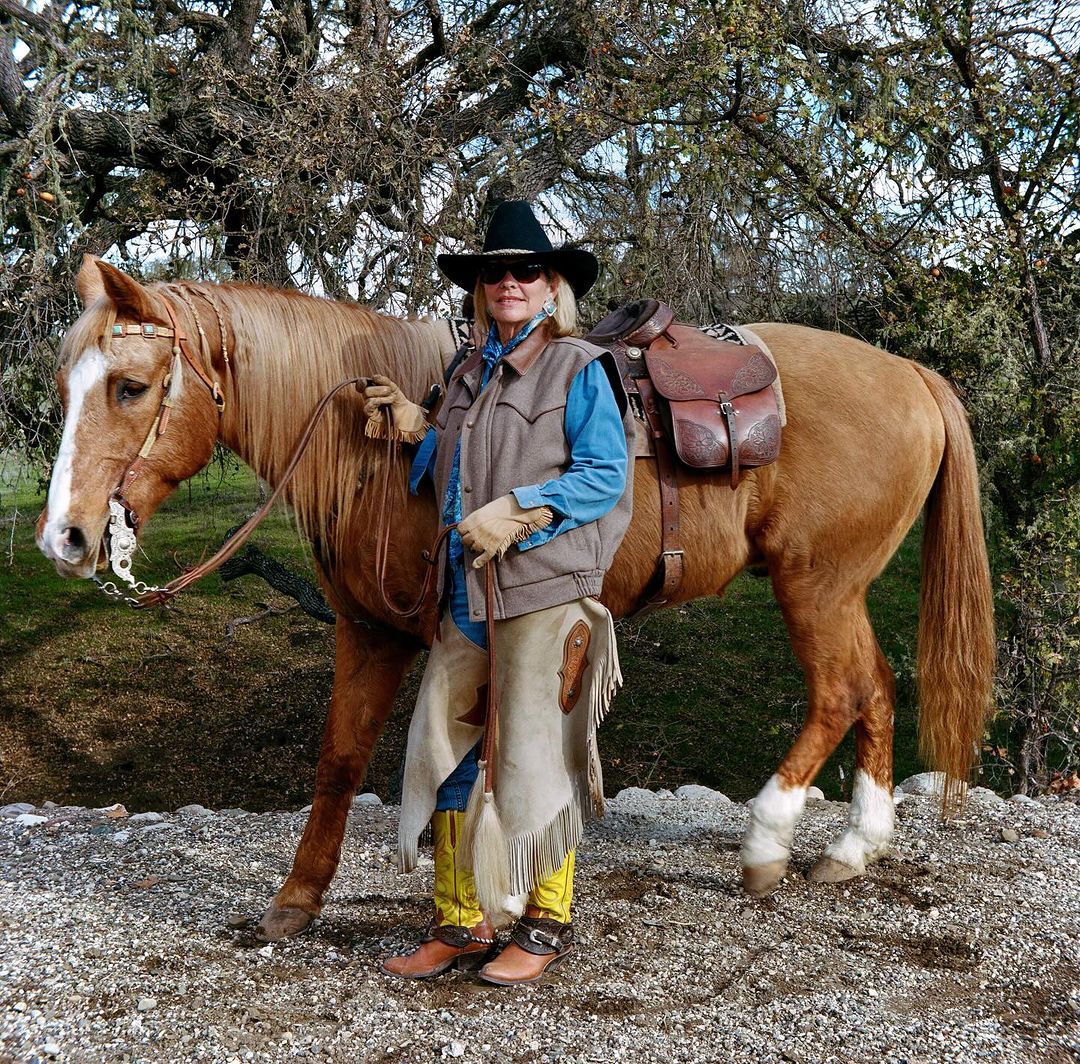 A woman in a cowboy hat and vest stands next to a palomino horse in an outdoor setting with trees and gravel ground.