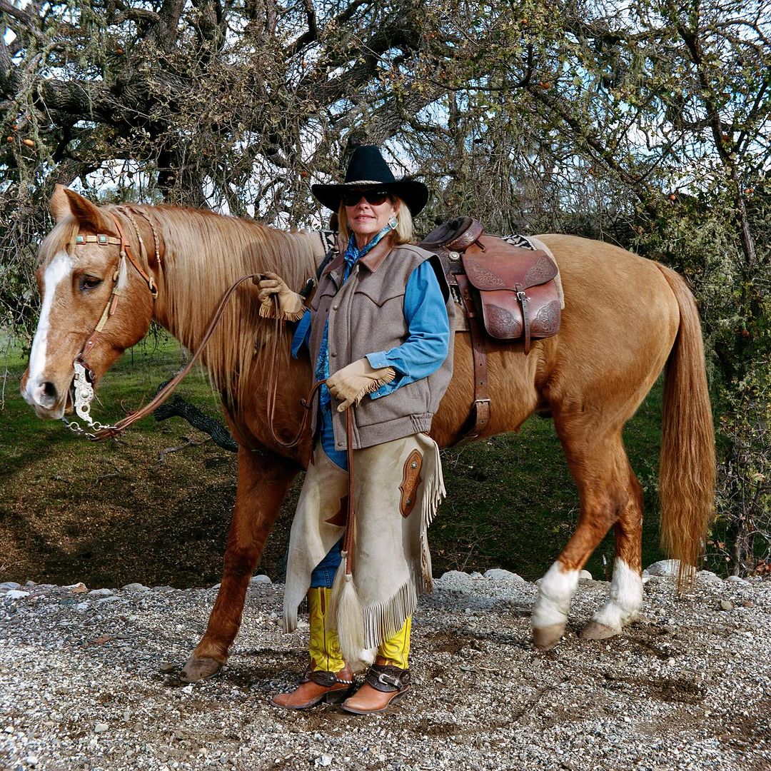 Woman poses with her horse outdoors, wearing chaps, vest, cowboy hat, and sunglasses, with trees in the background.
