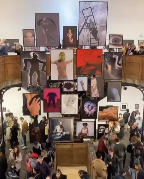 Art gallery exhibit featuring diverse framed artwork with people viewing the exhibition from above, creating a dynamic perspective.