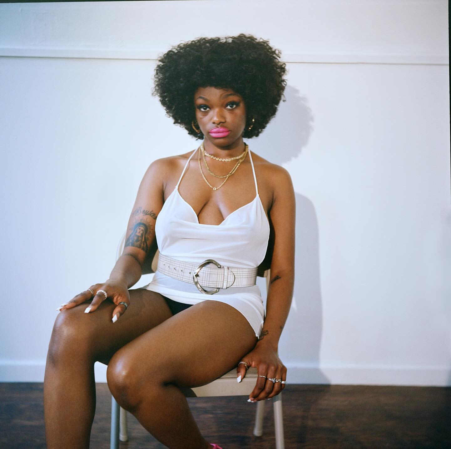 Portrait of African-American woman with afro wearing a white dress and chunky belt, sitting on a chair in front of a white wall.