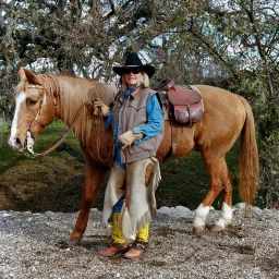 Woman in cowboy hat and vest standing next to a light brown horse outdoors under trees