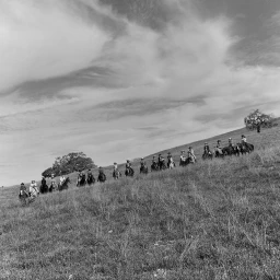 Black and white photograph of a line of equestrians riding horses across a sloping grassy hillside under a dramatic cloudy sky.