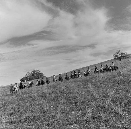 Group of horseback riders traversing a grassy hillside under an expansive overcast sky in a black and white photograph.