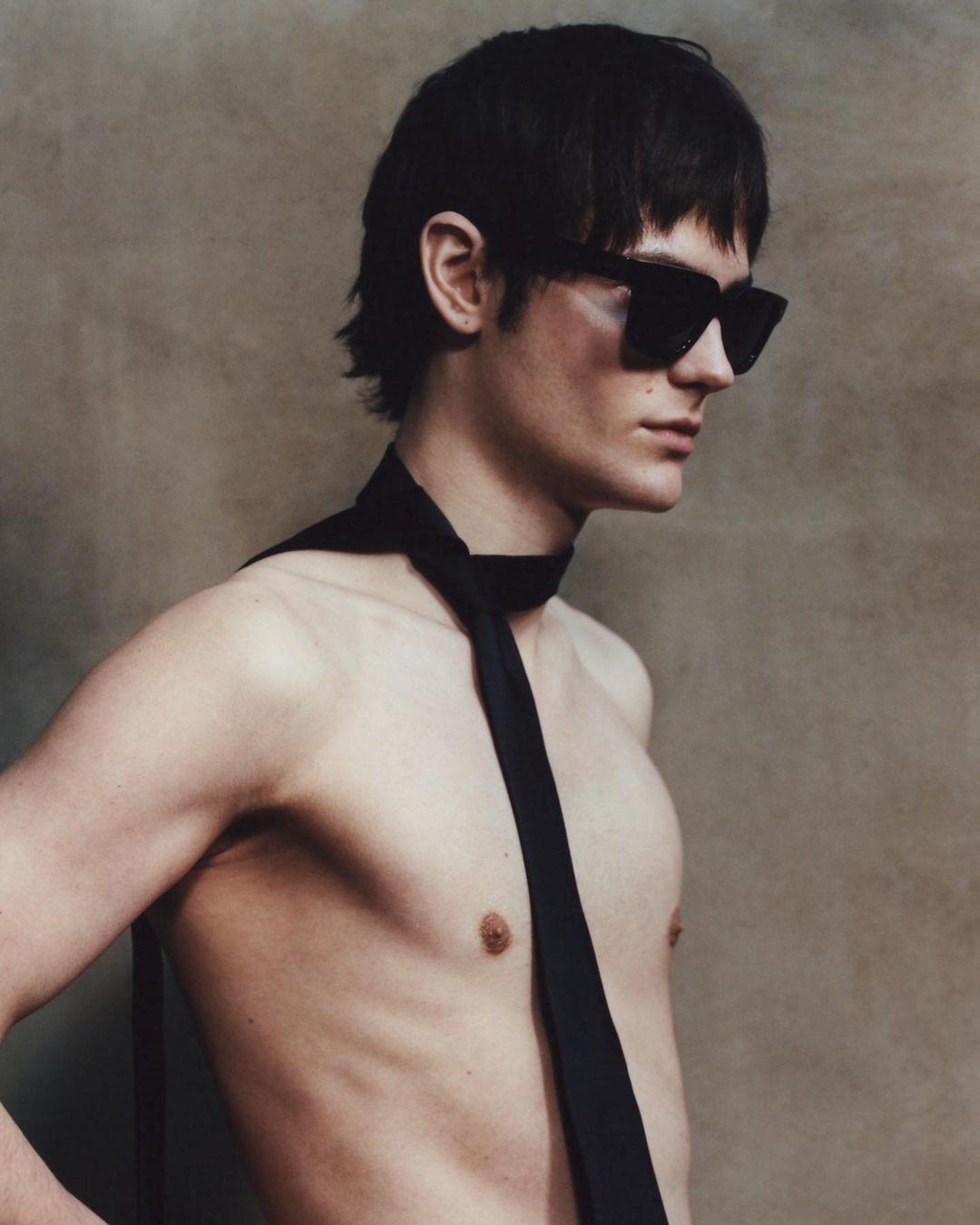 Striking close-up portrait of a shirtless adult male model with dark hair wearing sunglasses and a tie, set against a neutral backdrop.