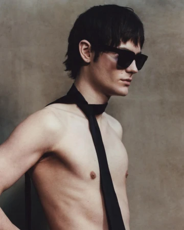 Striking close-up portrait of a shirtless adult male model with dark hair wearing sunglasses and a tie, set against a neutral backdrop.