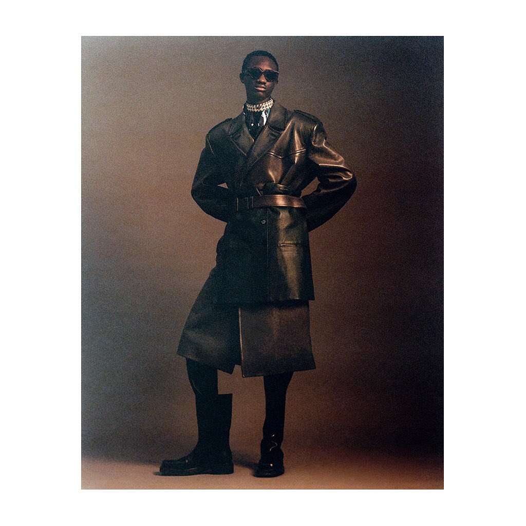 Stylish Black model in leather coat, skirt and boots poses in studio. Sunglasses and jewelry add to high fashion look.
