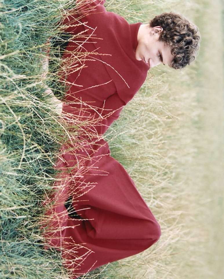Man in red robe lying in tall green grass. Relaxed male model with curly hair poses outside in a robe surrounded by natural grasses.