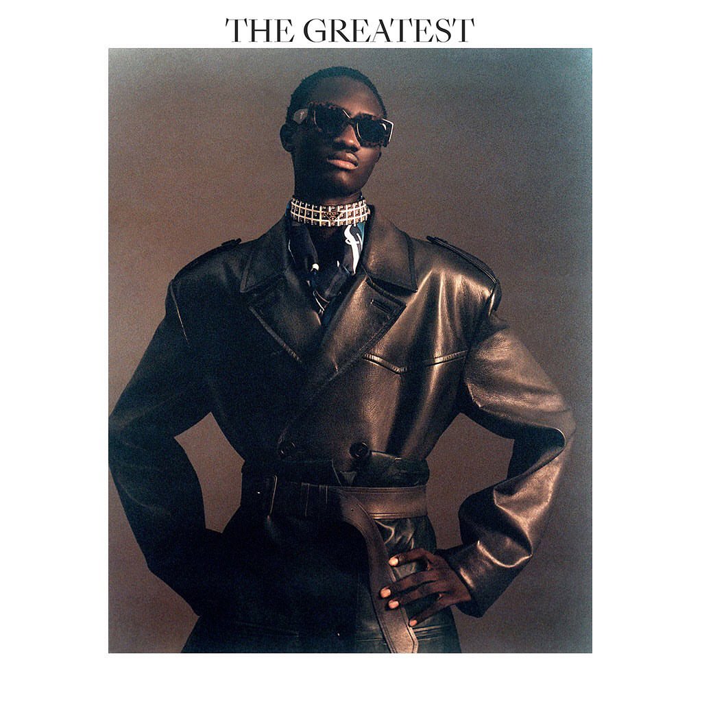 Portrait of an African American male model wearing a leather coat, neck jewelry and dark sunglasses, set against a neutral backdrop.