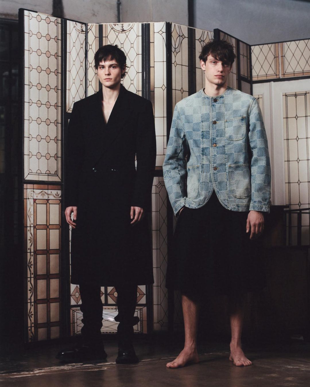 Two male models in a studio stand against a decorative screen; the model on the left wears a black coat, and the model on the right wears a denim jacket.