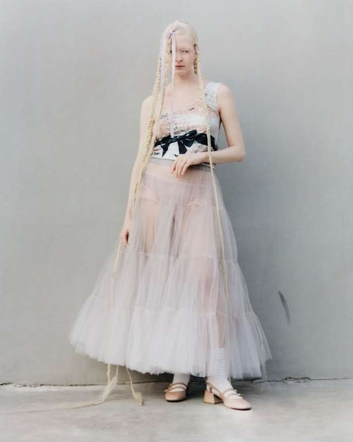 Albino woman with long blonde braids poses in a sheer white skirt and floral top against a grey wall, wearing pink shoes.