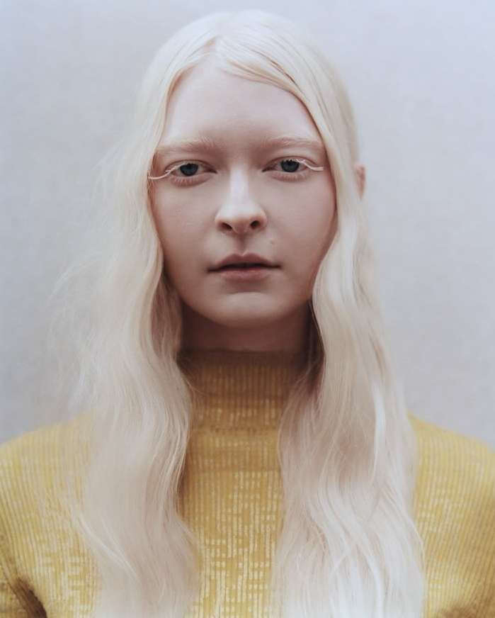 Close-up portrait of a woman with fair skin and hair, wearing a yellow sweater against a soft white background in a studio setting.