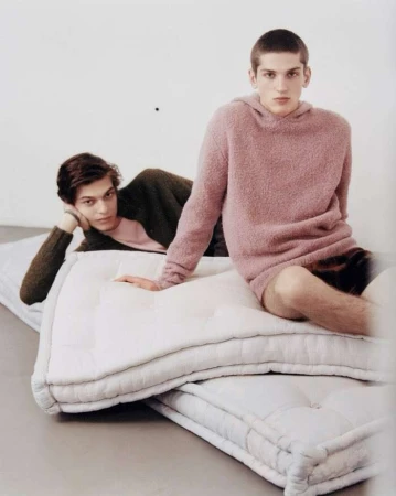 Two young adult males pose indoors on stacked white quilted mattresses, one seated, one reclining, wearing knit sweaters.